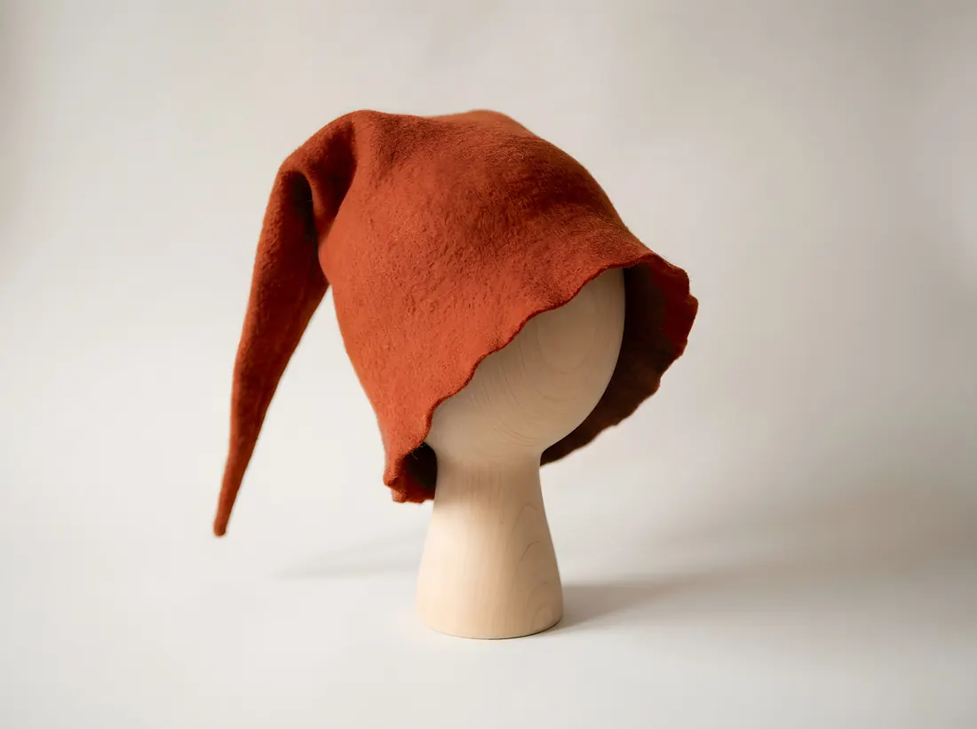 A soft pointed felt hat in rust red on a plain wooden stand, its long tip flopping over to one side
